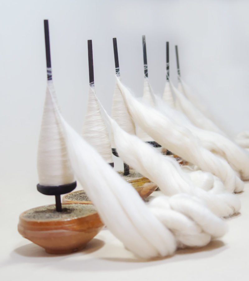 [Description: Six small wooden bowls lined up on a right diagonal angle leading from foreground to the white background, each with spun fabric on them. Near the base of the rods are threaded wooden bearings to hold the fabric in place.] 
