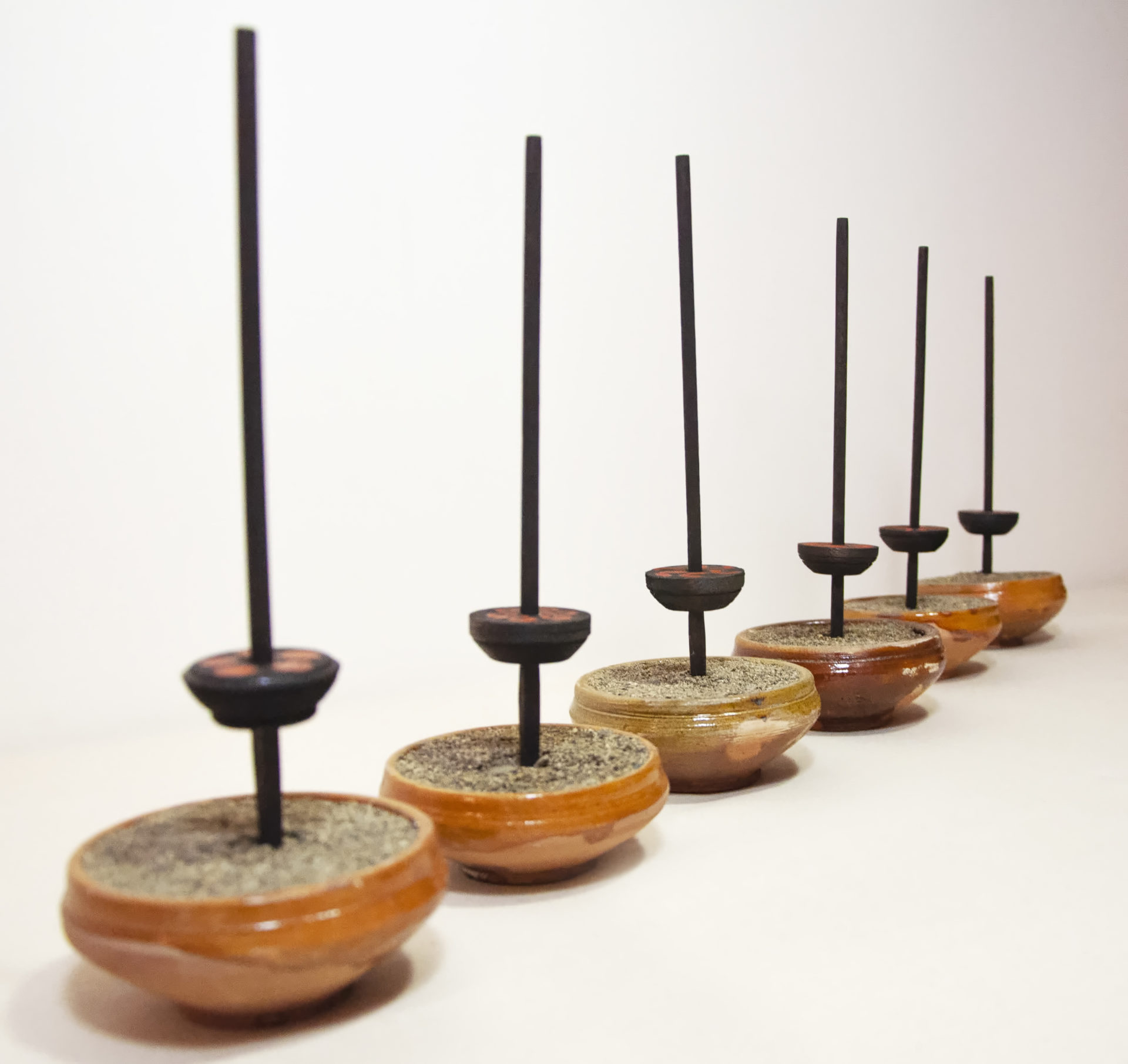 [Description: Six small wooden bowls lined up on a right diagonal angle leading from foreground to the white background, each filled with sand. One black, wooden rod in the center of each bowl. Near the base of these rods are threaded wooden bearings.] 