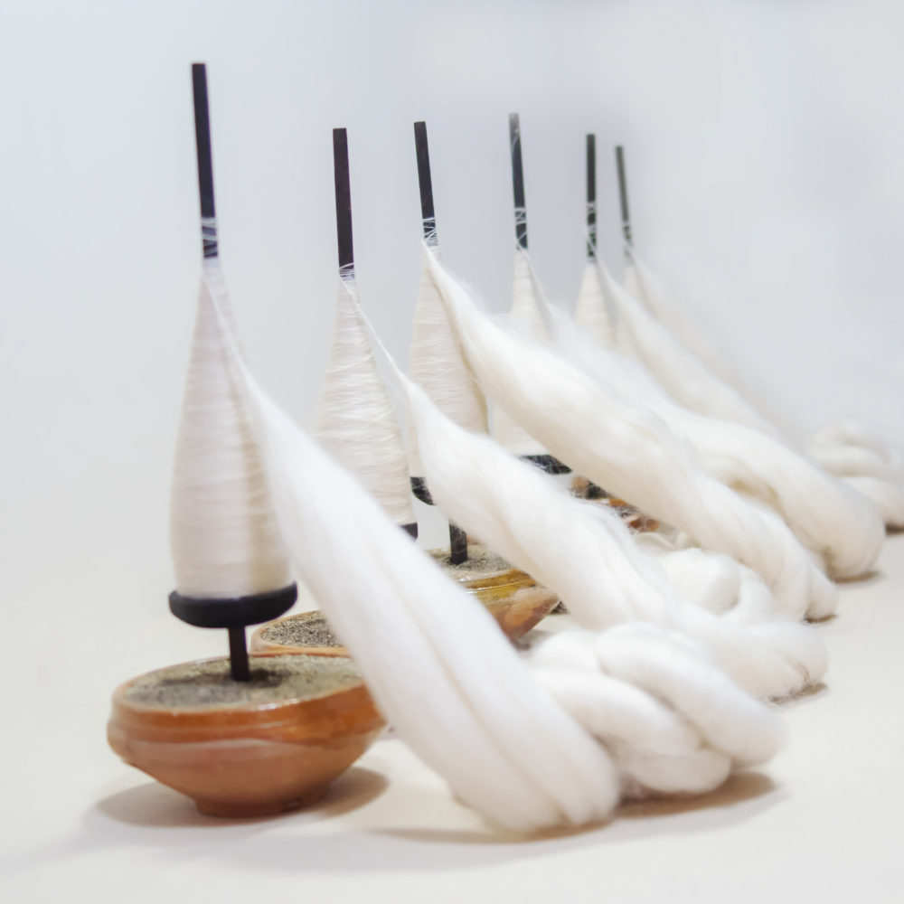 [Description: Six small wooden bowls lined up on a right diagonal angle leading from foreground to the white background, each with spun fabric on them. Near the base of the rods are threaded wooden bearings to hold the fabric in place.] 
